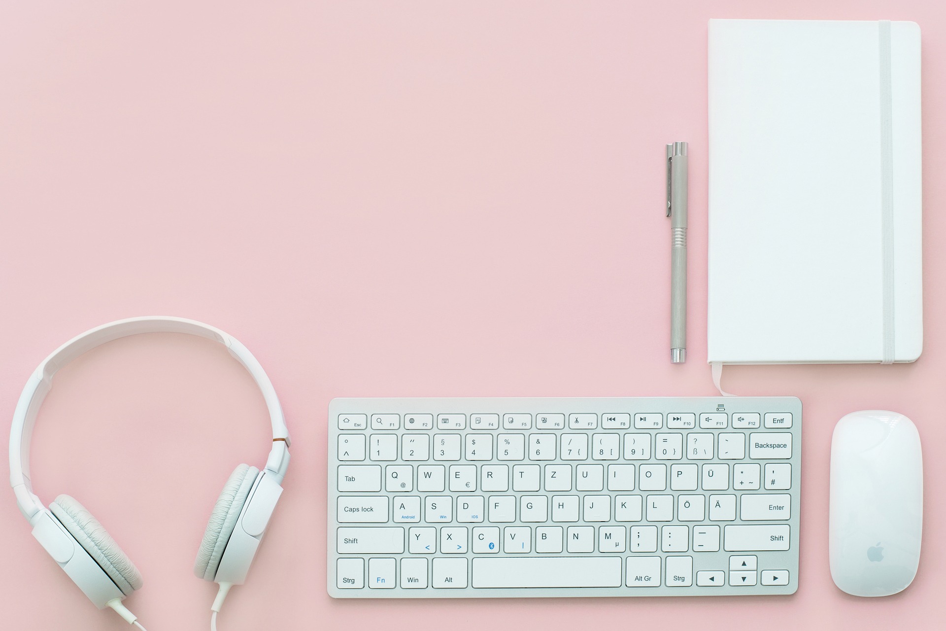 Une photo prise de haut d'un bureau rose avec un casque, un clavier, une souris, un bloc-note. Le tout est blanc.